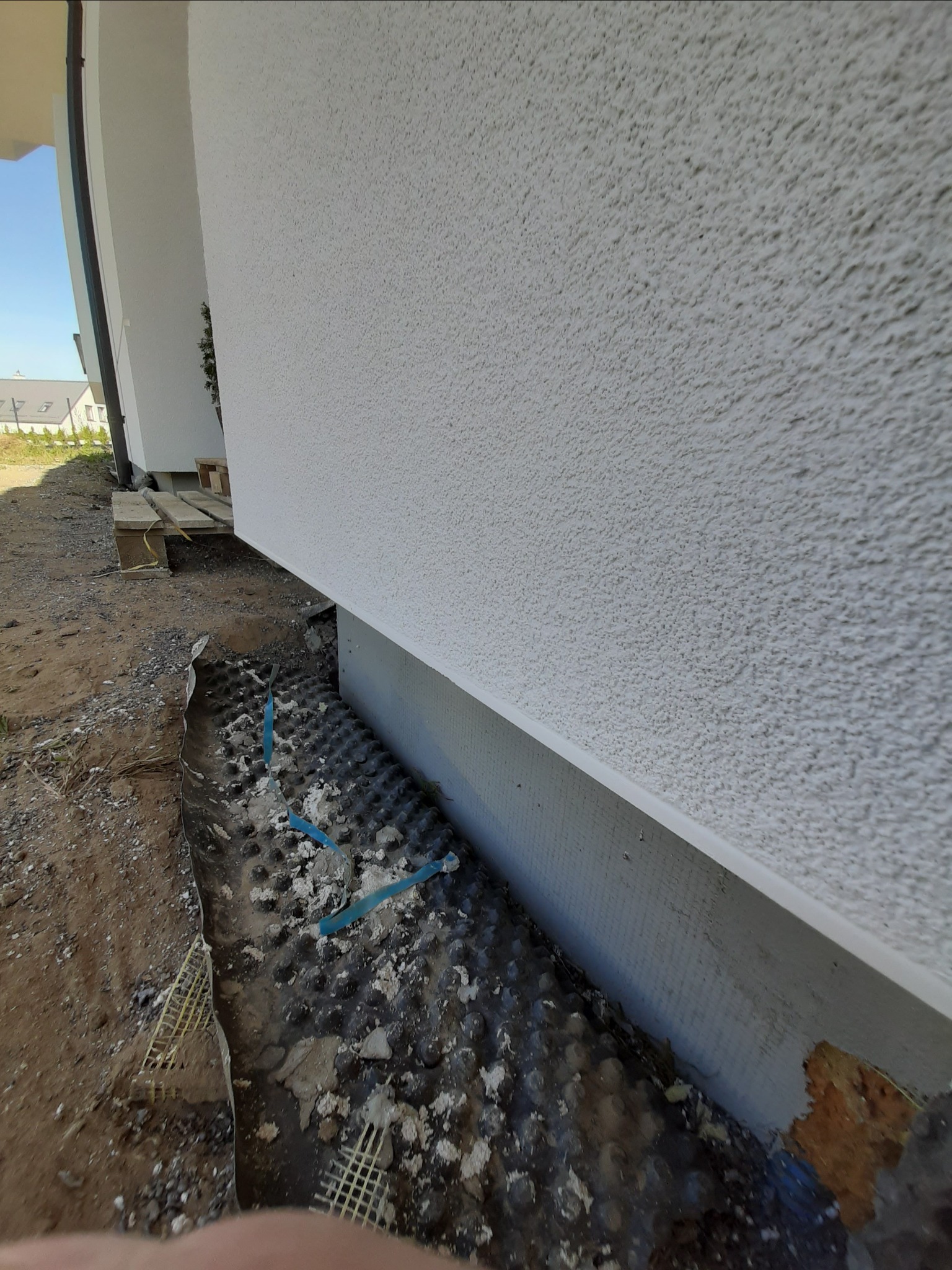 Ocieplanie fundamentów budynku z widoczną membraną kubełkową, izolacją termiczną i fragmentem elewacji o strukturze baranka.