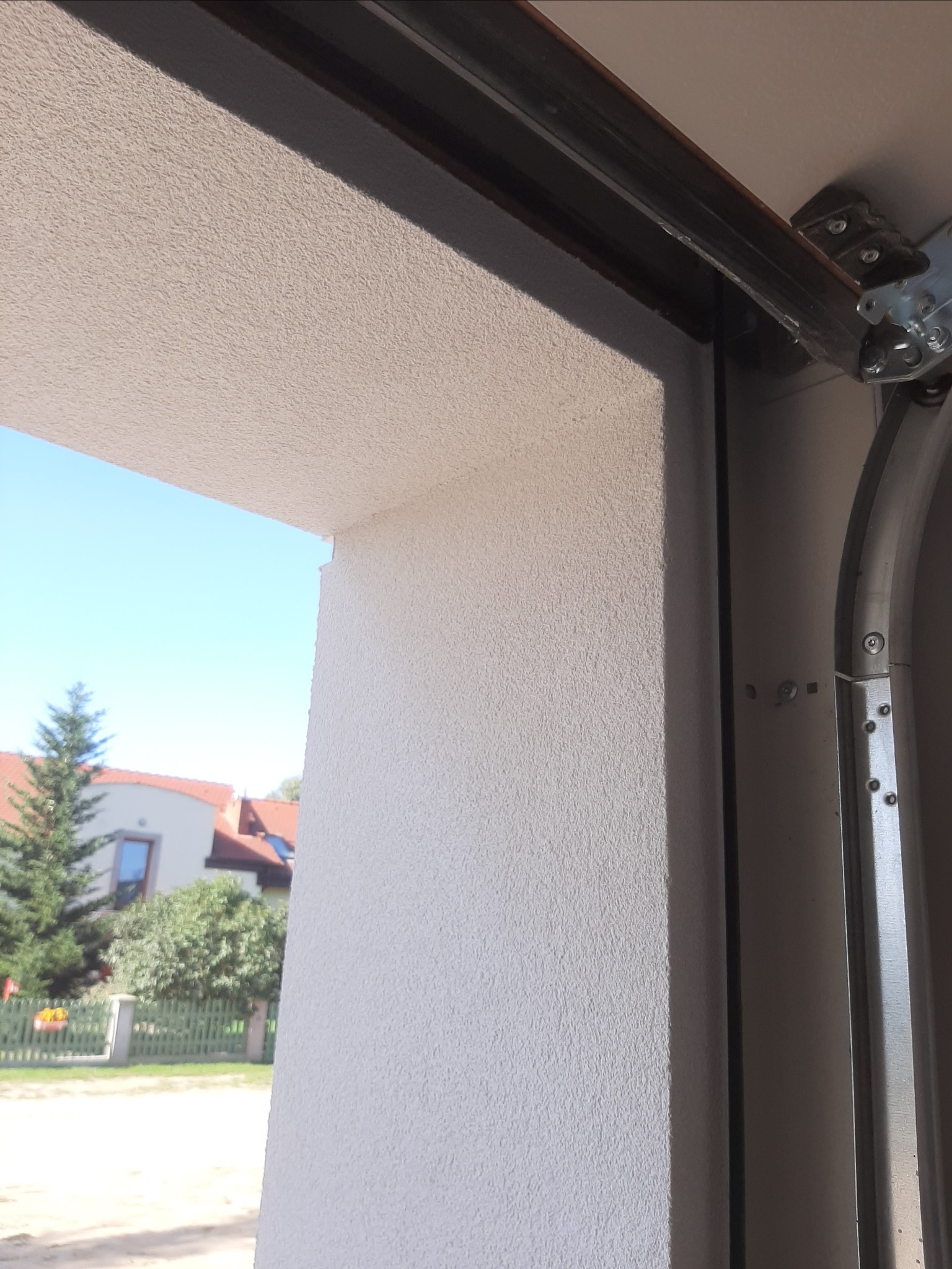 Ściana garażu z nową, jasną strukturą tynku baranka, widok z wnętrza garażu na zewnątrz, fragment mechanizmu otwierania bramy garażowej.
