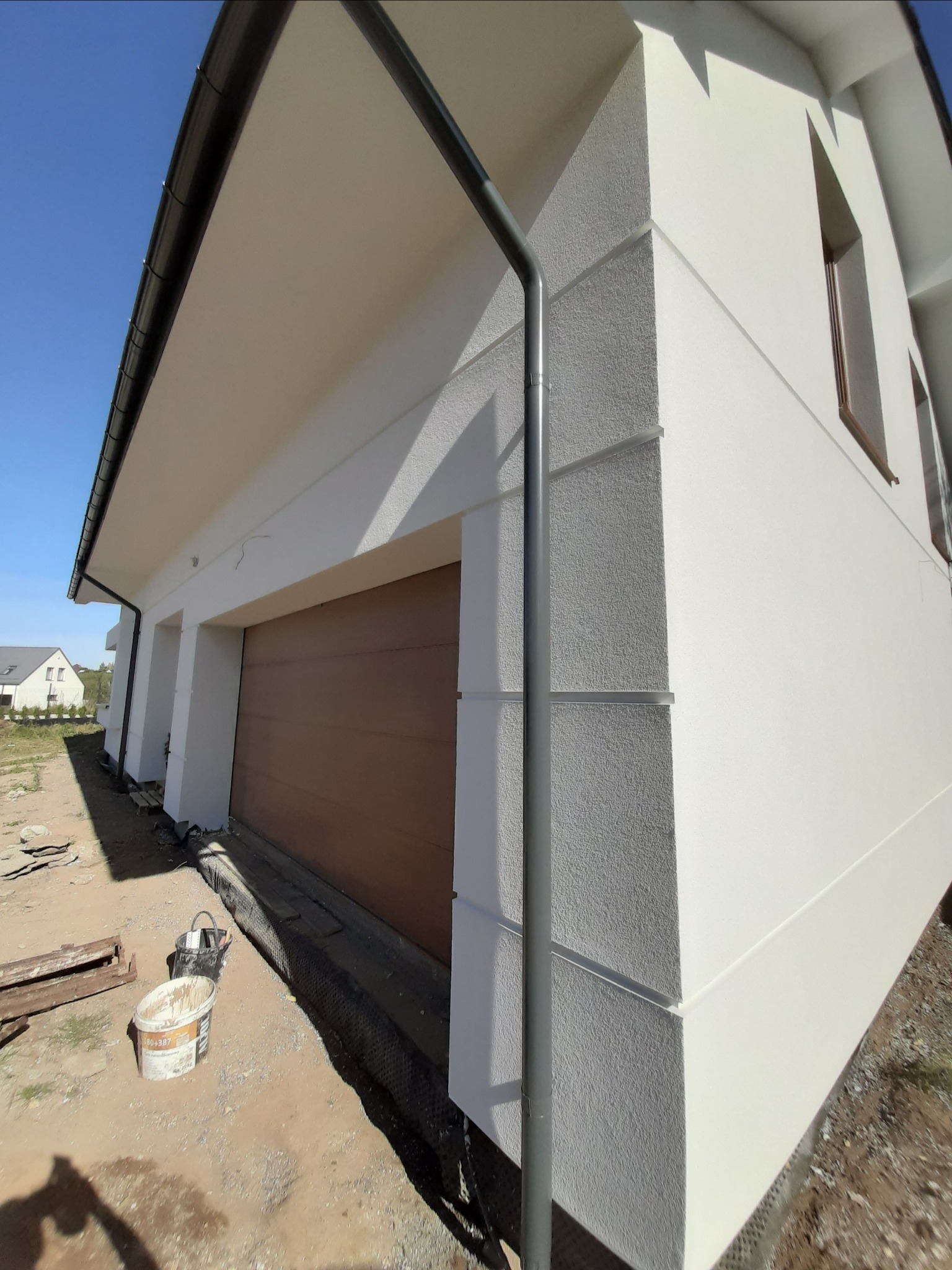 Świeżo otynkowana, biała elewacja domu z brązową bramą garażową, widoczny fragment rynny i przygotowane wiadro z zaprawą.