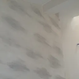 Ściana w rogu pomieszczenia z dekoracyjną, szarą strukturą na białym tle, widoczne fragmenty instalacji hydraulicznej.