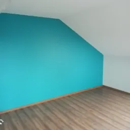 Pomalowane na turkusowo skośne poddasze z panelami podłogowymi, widok na róg pokoju z białym sufitem i fragmentem instalacji wentylacyjnej.