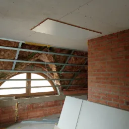 Poddasze w trakcie adaptacji: widoczne ocieplenie wełną mineralną między metalowymi profilami, częściowo zamontowane płyty na suficie, okrągłe okno w szczycie, fragment ściany z czerwonej cegły