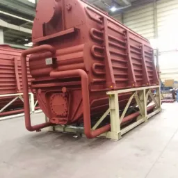 Duży, czerwony zbiornik przemysłowy z rurami i elementami mocującymi, umieszczony na beżowej metalowej konstrukcji wsporczej w hali produkcyjnej.