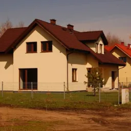 Osiedle nowych domów jednorodzinnych z brązowymi dachami i jasnymi elewacjami, ogrodzone siatką, na tle jesiennego krajobrazu.