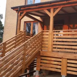 Zadaszona konstrukcja z drewna z schodami, prowadząca do podwyższonej części, z widocznymi elementami konstrukcyjnymi i balustradami, na tle budynku z oknami.
