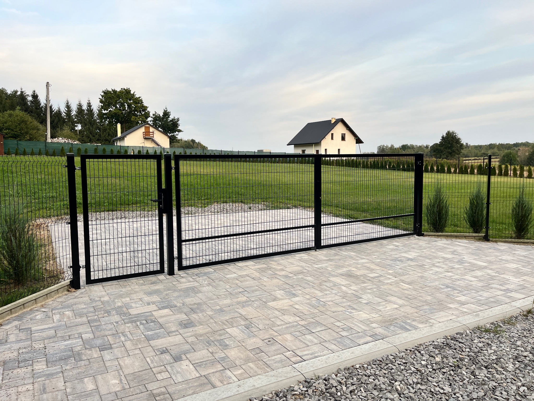 Dwuskrzydłowa, czarna brama wjazdowa z siatki na posesję z domem w tle, wyłożona kostką brukową w odcieniach szarości.