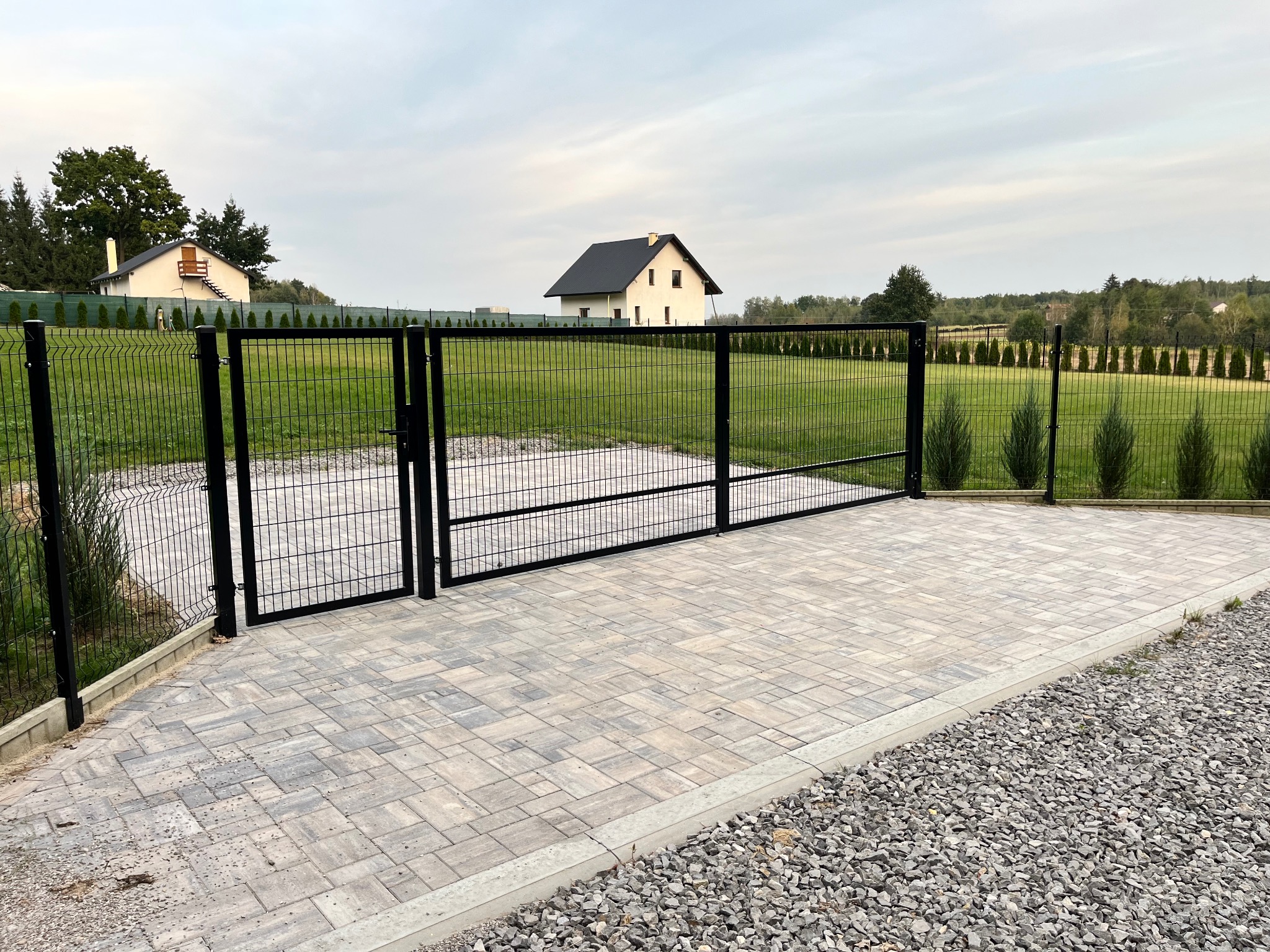 Nowoczesna, czarna brama dwuskrzydłowa z siatki panelowej, prowadząca na posesję z kostką brukową w odcieniach szarości, w tle dom jednorodzinny i rząd tuj.