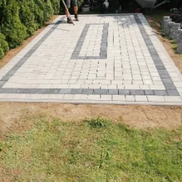 Człowiek z łopatą wyrównuje powierzchnię z kostki brukowej w jasnych i ciemnych odcieniach szarości, ułożonej we wzór z obramowaniem, na podwórku z tujami w tle.