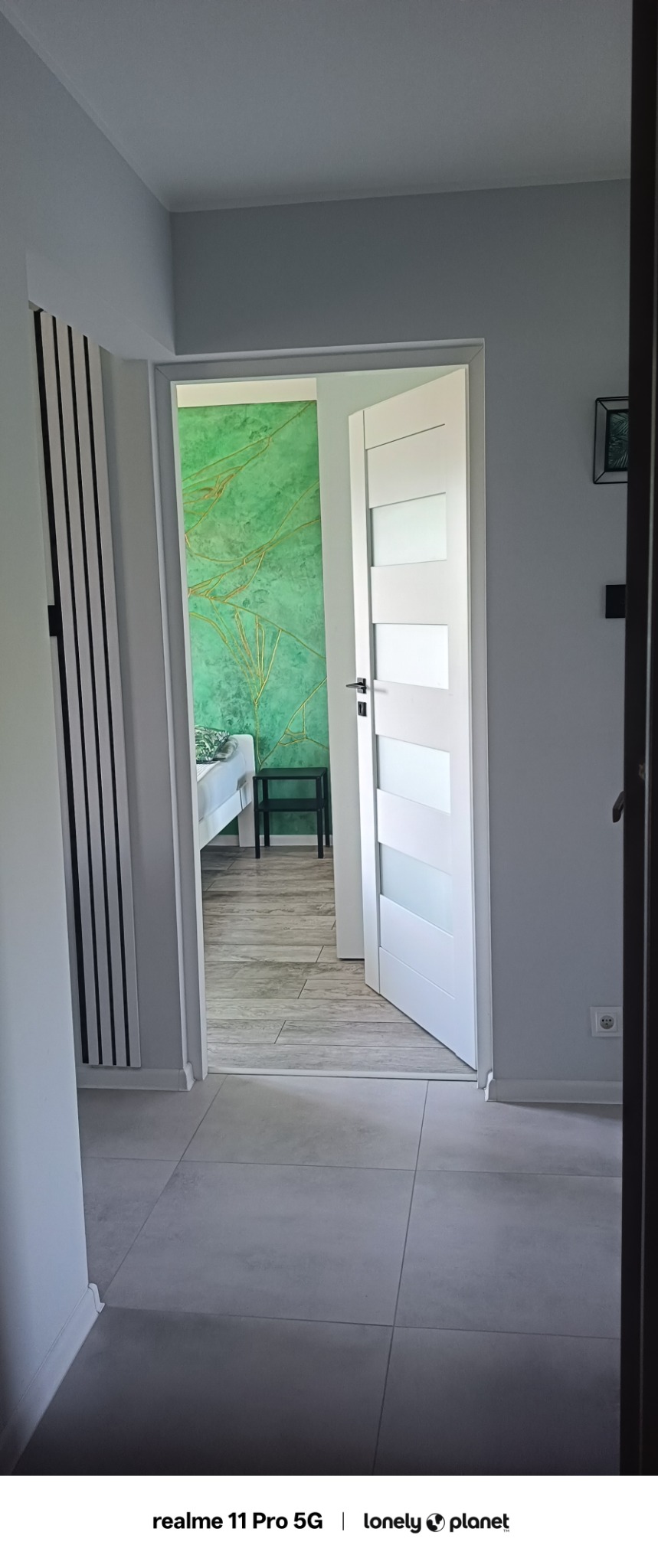 Wnętrze mieszkania z otwartymi białymi drzwiami. Widoczna zielona ściana z dekoracyjnym wzorem, szare płytki podłogowe i grzejnik panelowy.