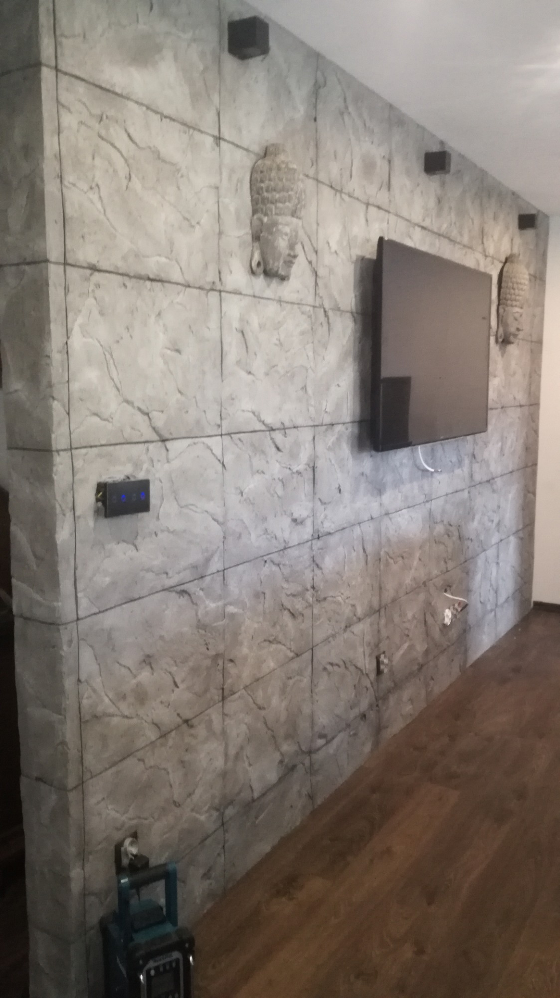 Ściana wykończona płytkami imitującymi kamień z dekoracyjnymi rzeźbami głowy Buddy, wiszący telewizor i drewniana podłoga, widoczny włącznik światła i narzędzie budowlane w rogu.