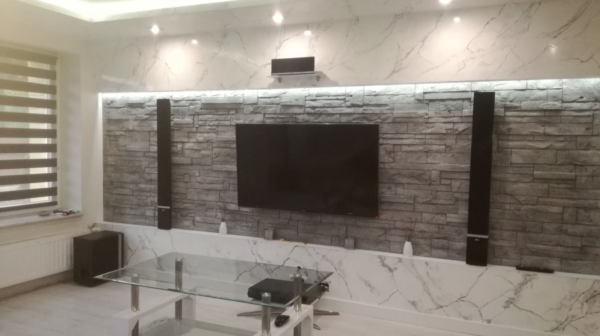 Wykończony salon z marmurową ścianą i ceglanym panelem pod telewizorem, oświetlenie LED, głośniki kolumnowe i szklany stolik kawowy.
