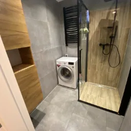 Nowoczesna aranżacja łazienki w Gryfinie: szare płytki, kabina prysznicowa typu walk-in z czarnymi profilami, drewniane szafki i pralka.