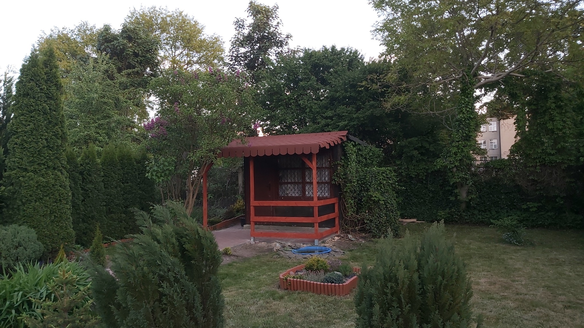 Drewniana altanka z czerwonym dachem i obudową, otoczona zielenią drzew i krzewów w ogrodzie, widok na budynek w tle.