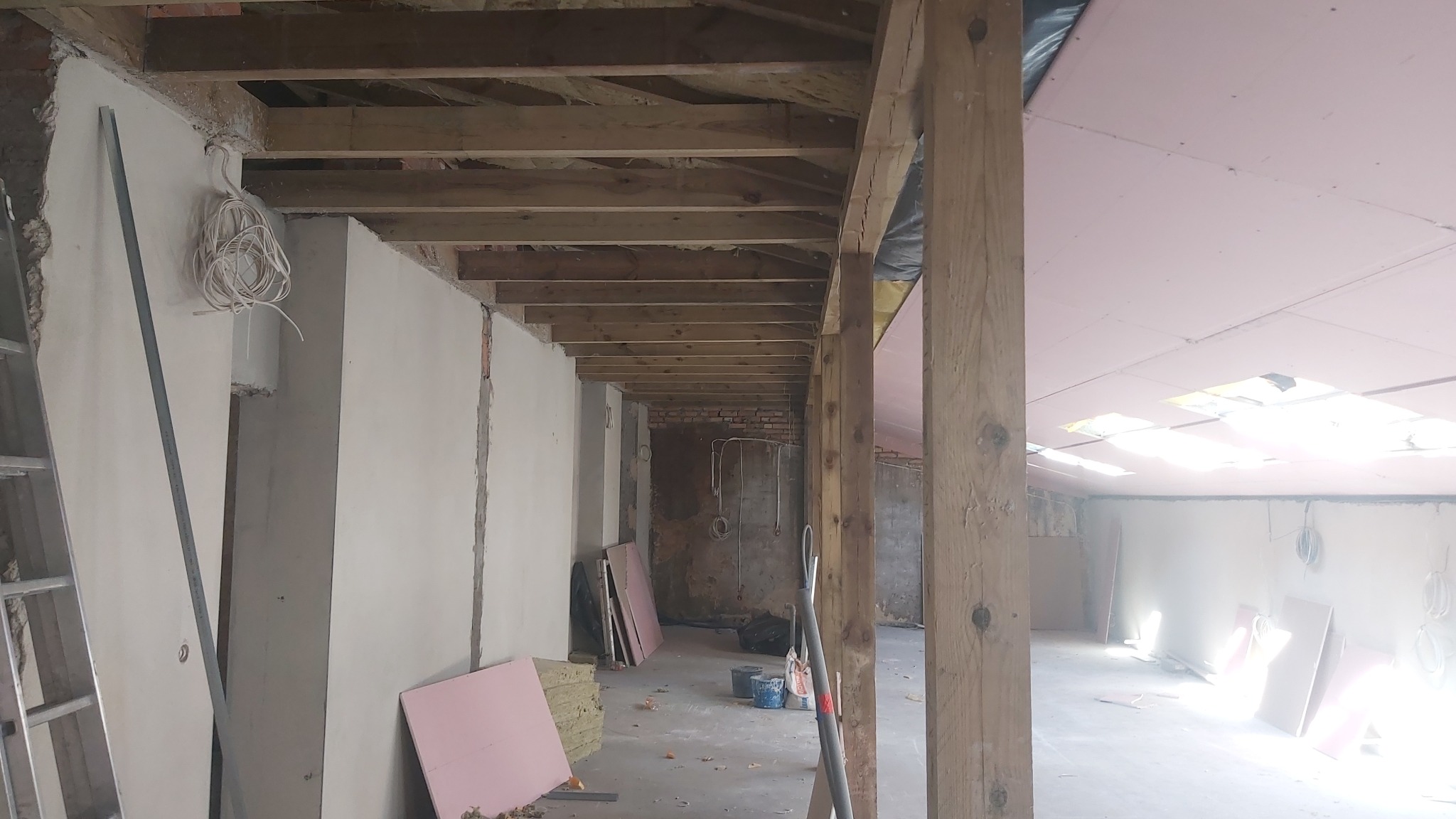 Wnętrze w trakcie remontu: surowe ściany, drewniane belki stropowe, częściowo zamontowana płyta karton-gips na suficie, widoczne kable elektryczne i drabina oparta o ścianę.