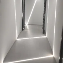 Nowoczesny korytarz z białymi ścianami i podłogą, oświetlony liniowymi lampami LED tworzącymi geometryczny wzór na ścianach i suficie, z otwartymi drzwiami w głębi.