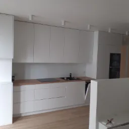 Nowoczesna, minimalistyczna kuchnia z białymi frontami szafek, drewnianym blatem i oświetleniem punktowym na suficie.