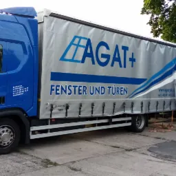 Ciężarówka firmy Agatplus z reklamą okien i drzwi, zaparkowana na betonowym placu.