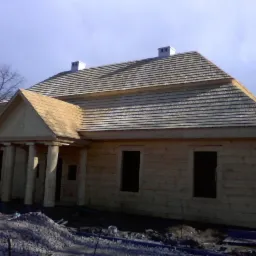 Nowy dom z drewnianych bali z dachem pokrytym gontem drewnianym, widoczne kolumny wejściowe i kominy, teren wokół w trakcie prac wykończeniowych.