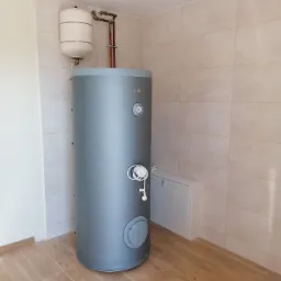 Szary piec CO z białym zbiornikiem wyrównawczym i miedzianymi rurami w nowym pomieszczeniu z beżowymi płytkami na ścianach i drewnopodobnymi płytkami na podłodze.