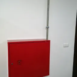 Czerwona szafka hydrantowa z kluczem na białej ścianie, obok pionowa rura instalacyjna z obejmą i włącznik światła.