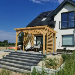 Drewniana konstrukcja z lamelami przy białym domu z panelami solarnymi, widok z dołu na taras z szarych płyt, z hamakiem i meblami ogrodowymi, niezagospodarowany teren wokół.