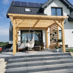 Drewniana konstrukcja z jasnego drewna na tarasie z meblami wypoczynkowymi, hamakiem i dekoracyjną ścianką z roślinami w doniczkach, na tle nowoczesnego domu z panelami słonecznymi.