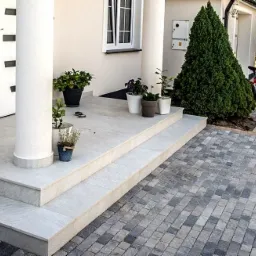 Schody wejściowe wyłożone jasnoszarą płytką, donice z kwiatami na podeście, kostka brukowa na podjeździe, czerwony rower oparty o tuję.