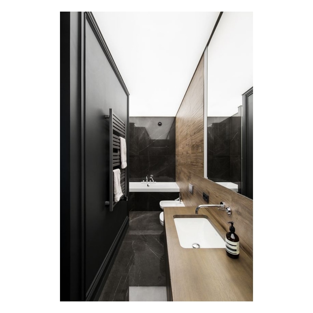 Nowoczesna łazienka z podświetlanym sufitem, czarnymi ścianami i marmurową podłogą, wyposażona w długi drewniany blat z umywalką, grzejnik drabinkowy i wannę w tle.