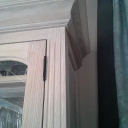 Detal wykończenia górnej części przeszklonej szafy z drewna o jasnym, bielonym wykończeniu, widoczny zawias drzwi i fragment zasłony w tle.