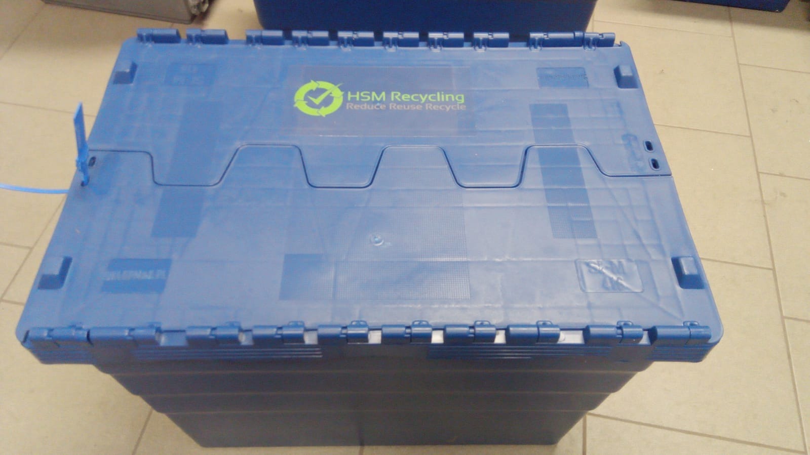 Niebieski, plastikowy pojemnik z logo HSM Recycling na jasnej podłodze z płytek.