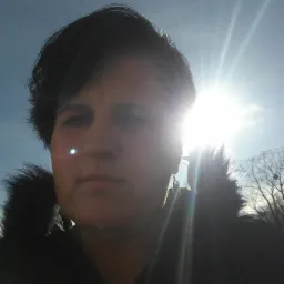 Portret osoby w kurtce z futrzanym kołnierzem, oświetlonej silnym światłem słonecznym, z widocznym kolczykiem i piercingiem w nosie.