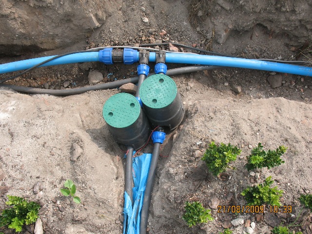 Instalacja automatycznego systemu nawadniania ogrodowego: widoczne podziemne elementy z zielonymi pokrywami, niebieskie rury doprowadzające wodę oraz kable elektryczne w wykopie ziemnym.