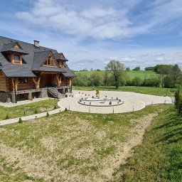 NOWBRUK - kompleksowe usługi brukarskie - Rozległy widok na drewniany dom z kamienną podmurówką, okrągły podjazd z kostki brukowej i fontanną, oraz wijącą się drogę w tle, w słoneczny dzień.