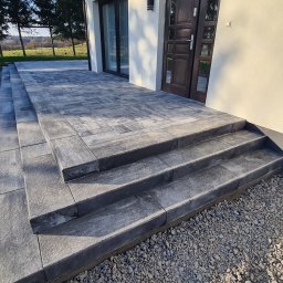 NOWBRUK - kompleksowe usługi brukarskie - Schody wejściowe i taras z kostki brukowej w odcieniach szarości, prowadzące do domu z ciemnymi drzwiami. Widoczne detale ułożenia kostki i otoczenie domu.