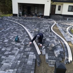 NOWBRUK - kompleksowe usługi brukarskie - Pracownik w trakcie układania szarej kostki brukowej z białymi obrzeżami na podjeździe przed domem z numerem 98. Widoczne narzędzia i materiały budowlane.
