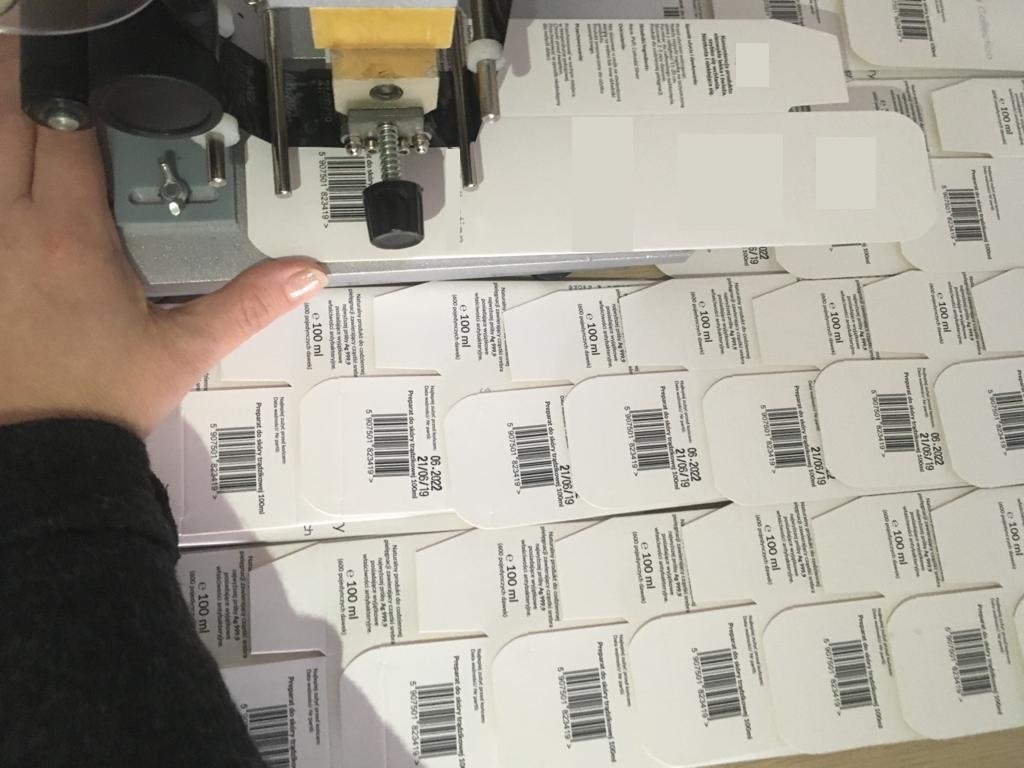 Automat etykietujący białe kartoniki z datą ważności, częściowo zasłonięte ręką, w tle stos opakowanych produktów.