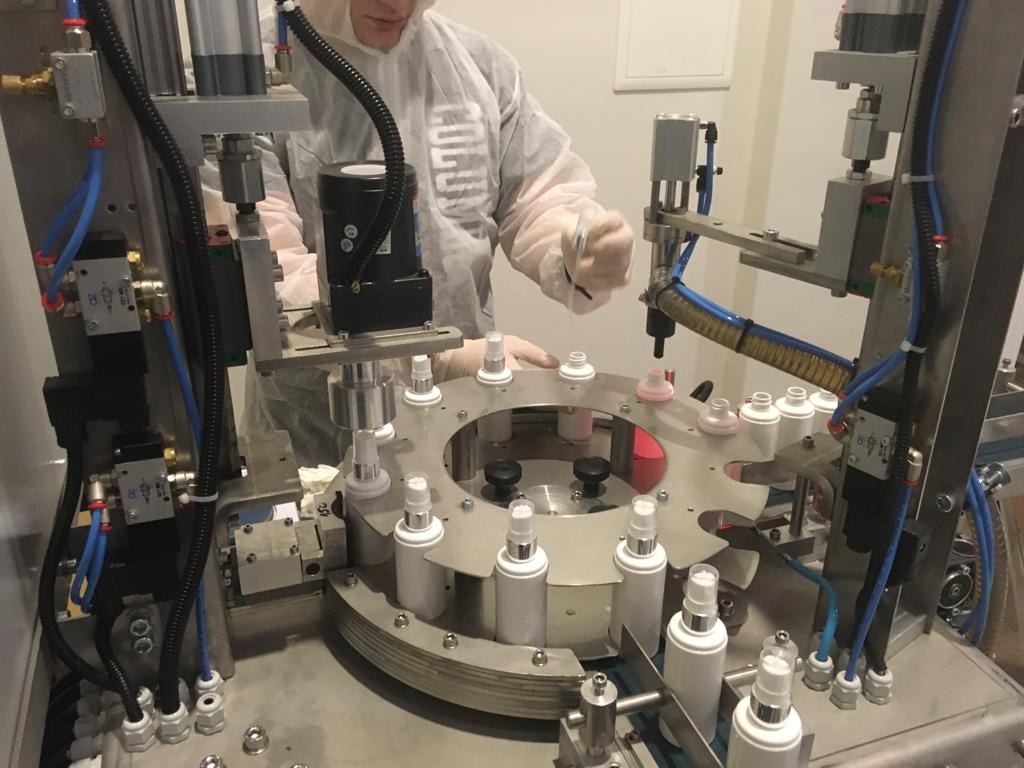 Automatyczna linia produkcyjna napełniająca białe butelki z atomizerem, obsługiwana przez osobę w kombinezonie ochronnym.