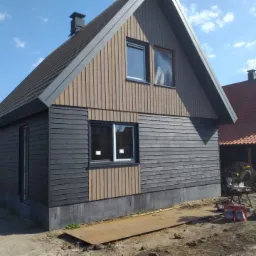 Nowoczesny dom z drewnianą elewacją w dwóch odcieniach szarości i brązu, widoczny fragment placu budowy z narzędziami.