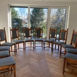 Ustawione w okręgu krzesła z drewnianymi ramami i szarymi siedziskami, po renowacji, na parkiecie ułożonym w jodełkę, widok przez okno na zewnątrz.