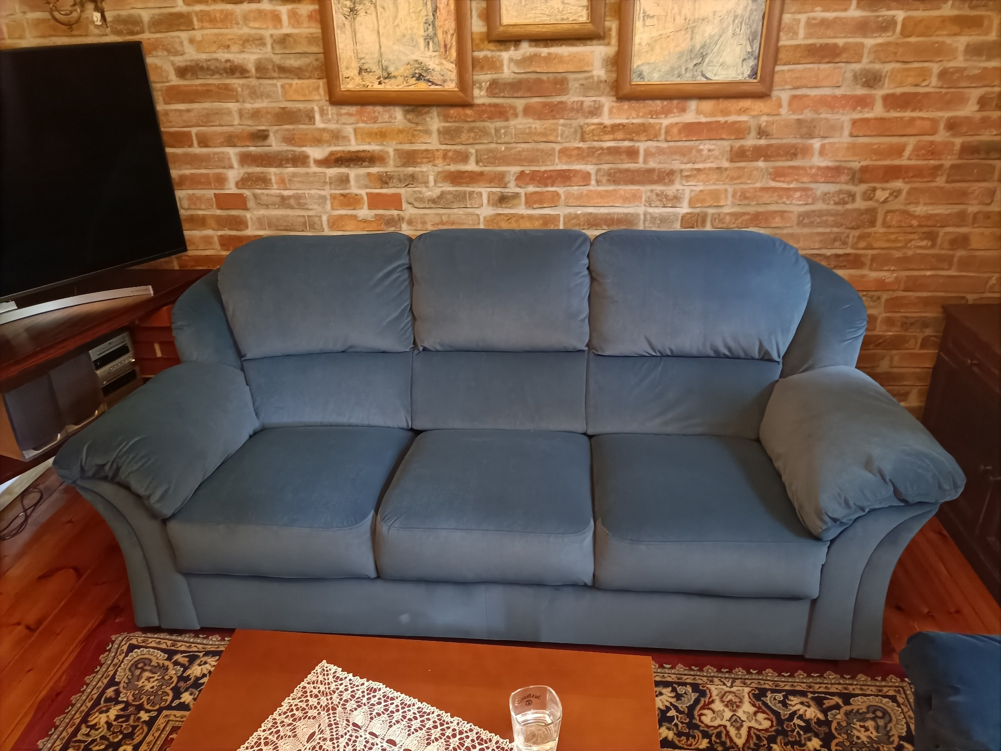 Trzyosobowa sofa w odcieniach niebieskiego, z welurową tapicerką, ustawiona na tle ściany z czerwonej cegły i drewnianej podłogi, z widocznym fragmentem stolika kawowego z koronkową serwetą...