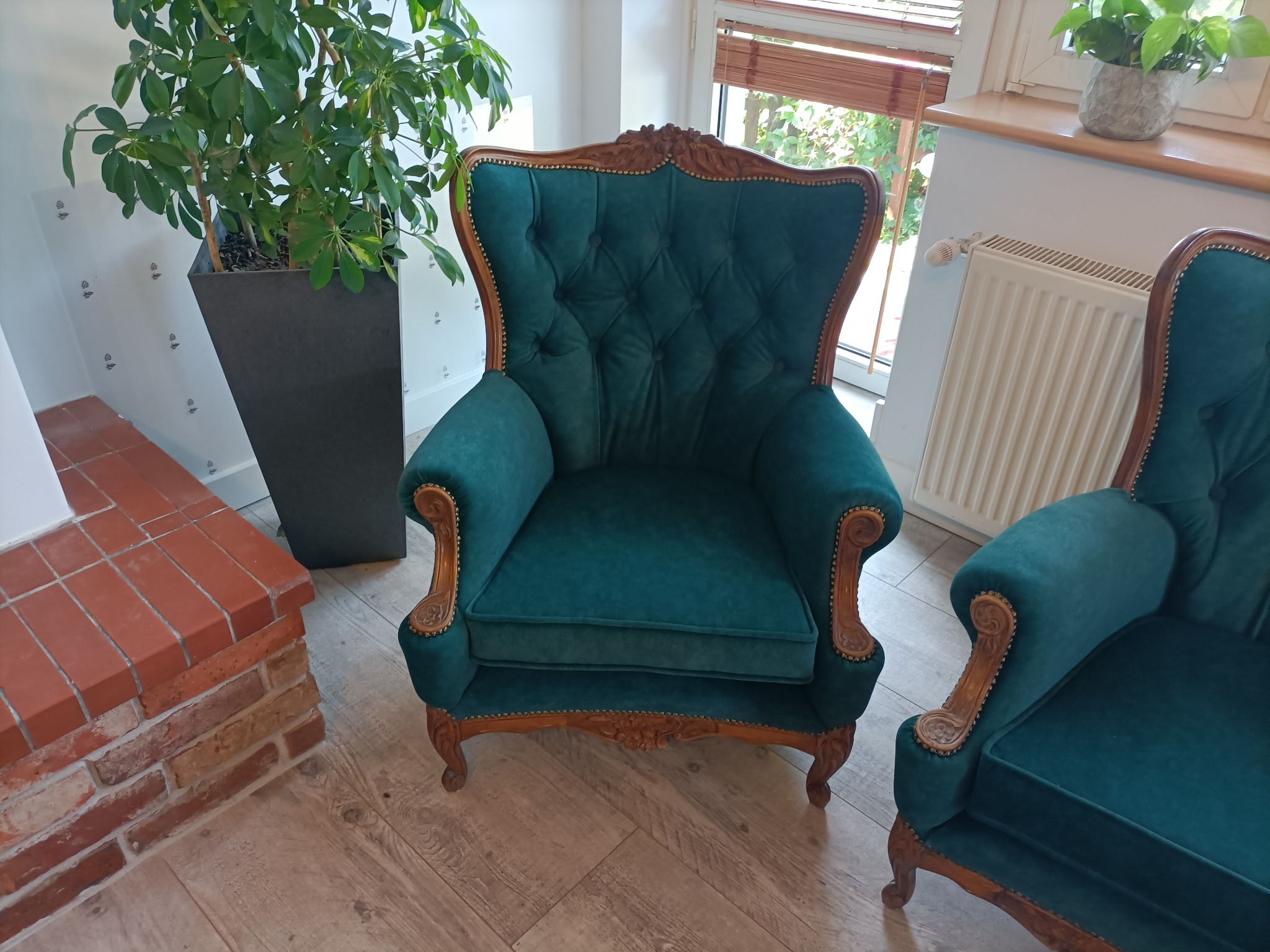 Dwa zielone, tapicerowane fotele w stylu vintage z drewnianymi zdobieniami, ustawione w jasnym wnętrzu z drewnianą podłogą i roślinami doniczkowymi.