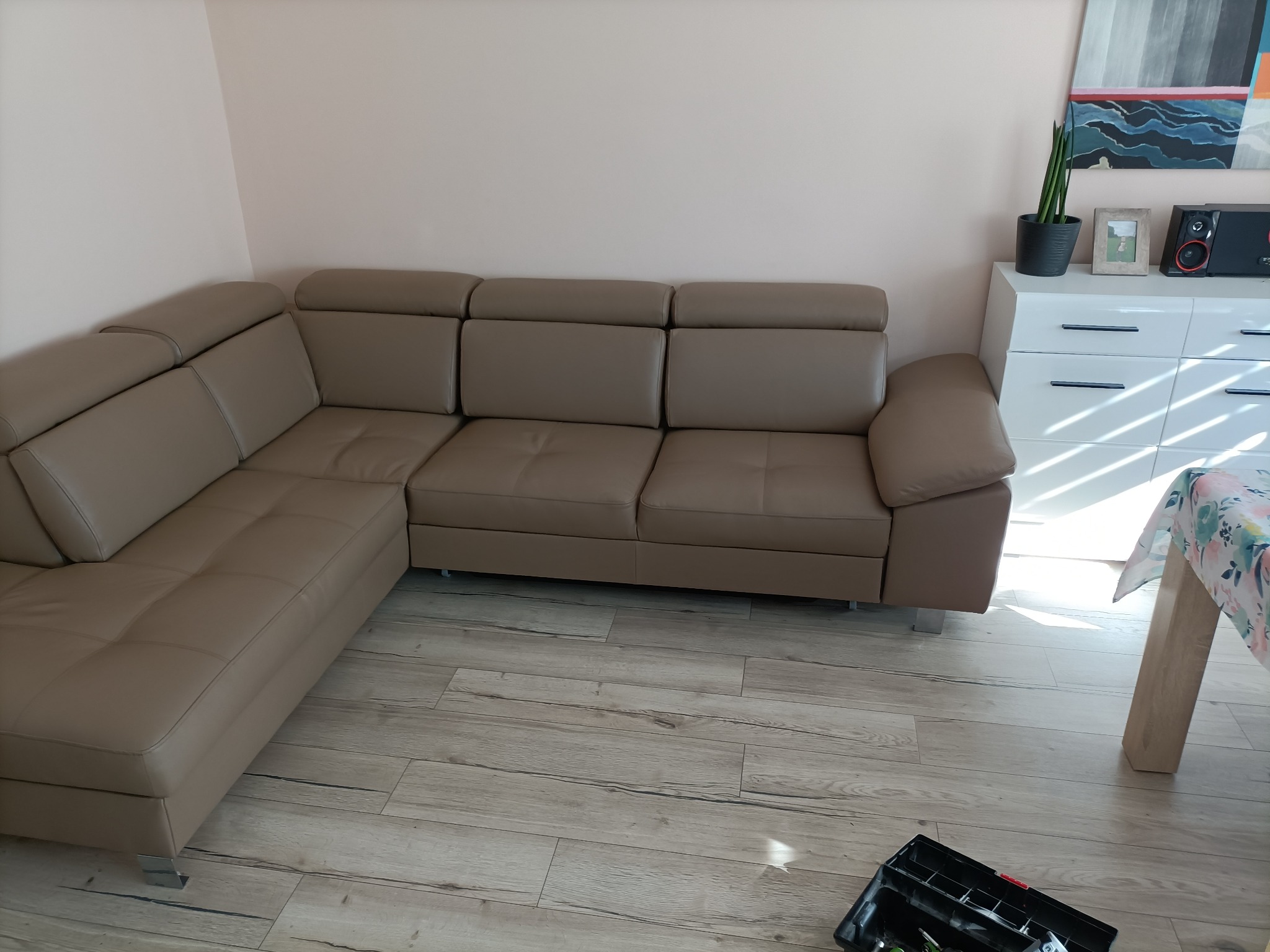 Beżowy narożnik z regulowanymi zagłówkami w salonie, widoczna skrzynka z narzędziami na podłodze, sugerująca montaż lub naprawę.