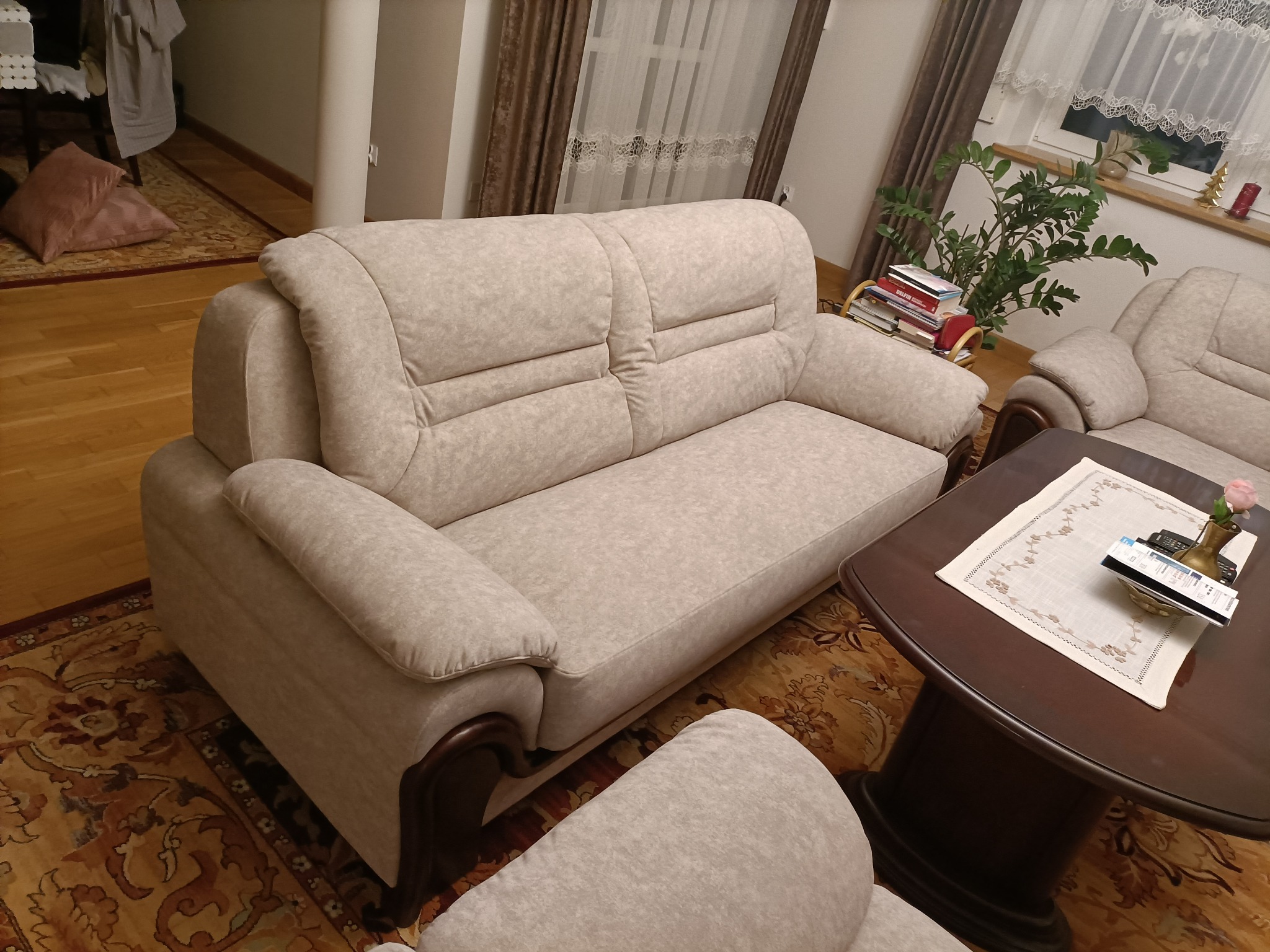Jasna, dwuosobowa sofa z drewnianymi elementami w salonie, widoczny fragment fotela i stolika kawowego z dekoracjami, dywan w orientalne wzory.
