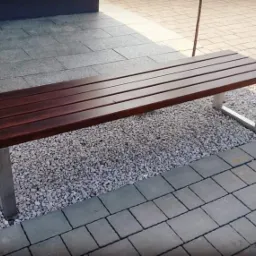 Ławka parkowa z drewnianym siedziskiem i metalowymi nogami w kształcie litery U, ustawiona na żwirowym podłożu, obok chodnika z szarych kostek.