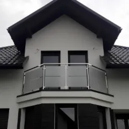 Balkon z balustradą ze szkła matowego i stali nierdzewnej, widok z dołu na tle pochmurnego nieba, czarny dach z dachówki ceramicznej.