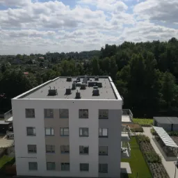 Nowoczesny apartamentowiec z płaskim dachem i licznymi kominami wentylacyjnymi, widziany z góry, na tle zieleni i błękitnego nieba z chmurami.