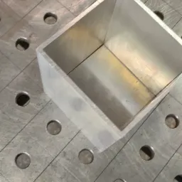 Aluminiowy prostopadłościan spawany na stole spawalniczym z otworami, widoczny ślad po spawaniu na krawędzi.