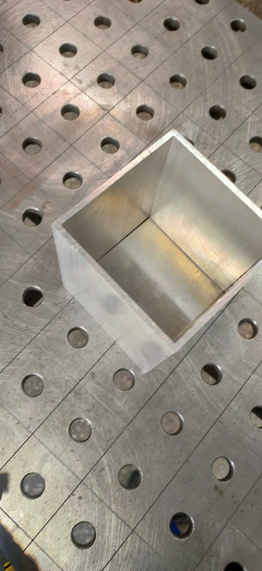 Aluminiowy prostopadłościan spawany na stole spawalniczym z otworami, widoczny ślad po spawaniu na krawędzi.