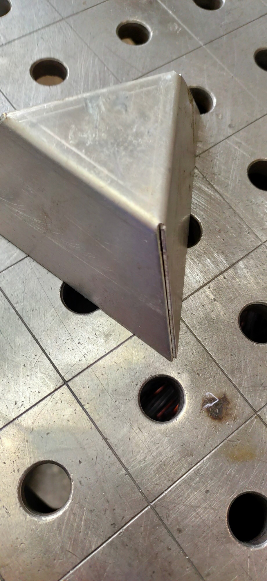 Metalowy trójkątny element spawany z blachy, umieszczony na stole spawalniczym z otworami, widoczne ślady obróbki i spawania.