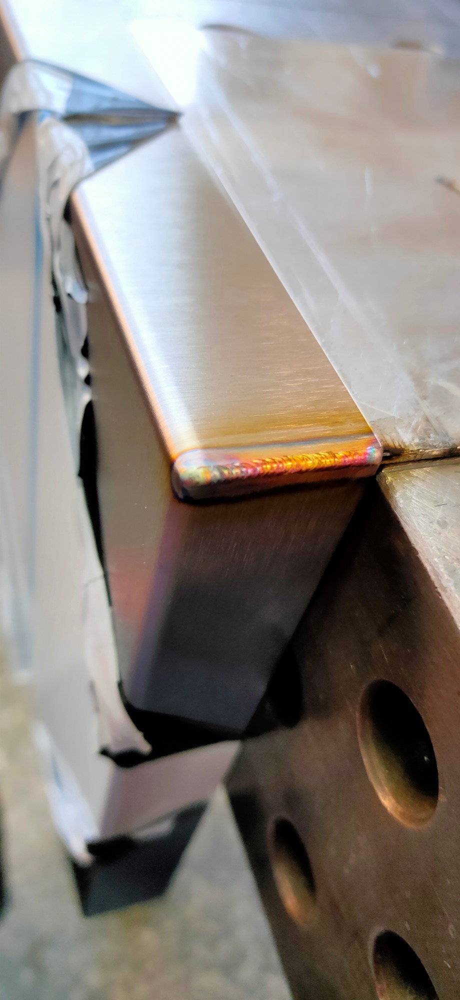 Zbliżenie na spawany narożnik metalowego profilu, widoczne tęczowe przebarwienia termiczne na spoinie, zabezpieczony folią.